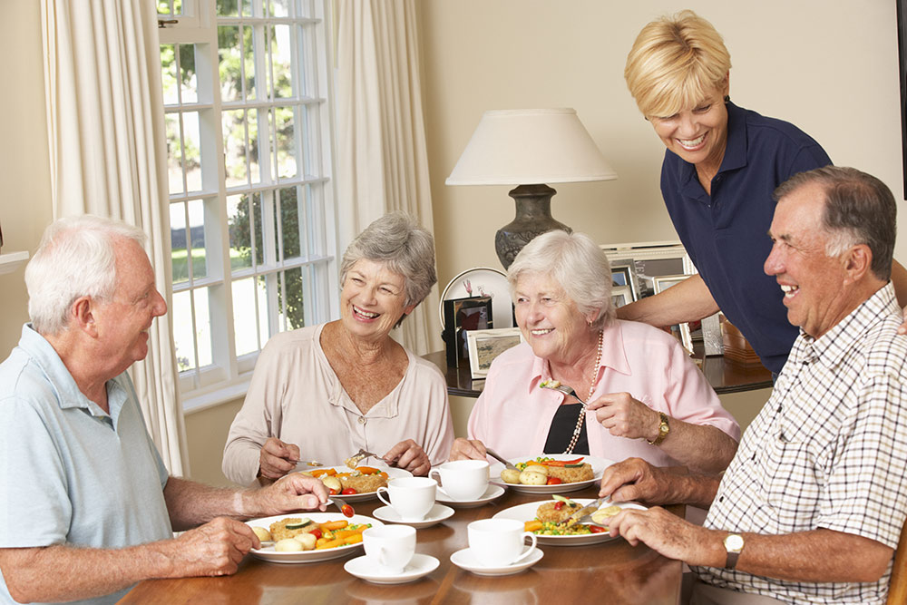 Happy smiling senior group eating meal at table with caregiver near by