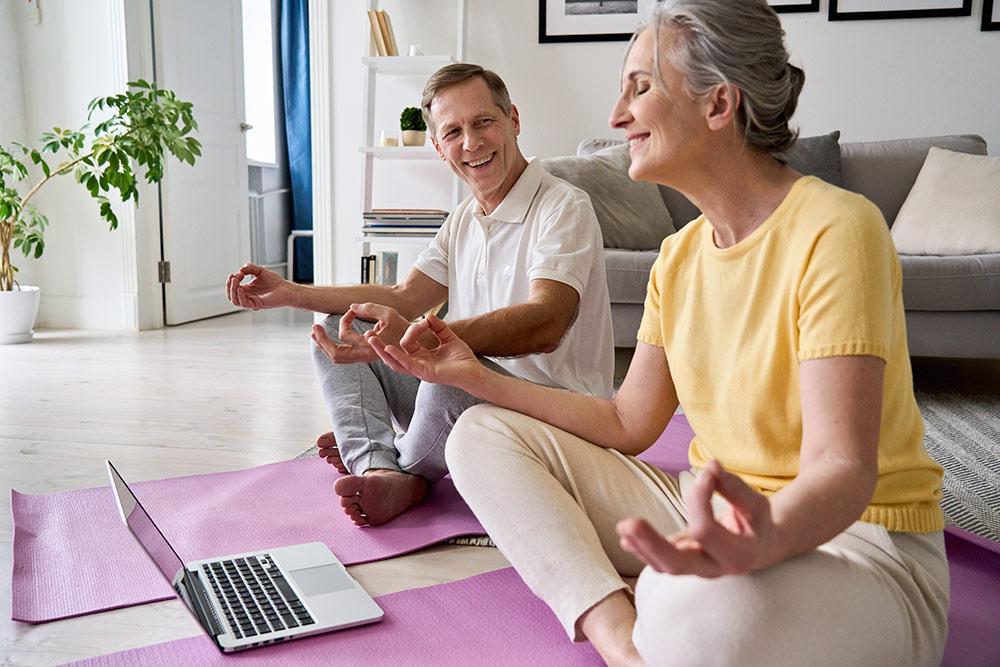 Senior couple sitting on floor in living room meditating during virtual meditation club on laptop