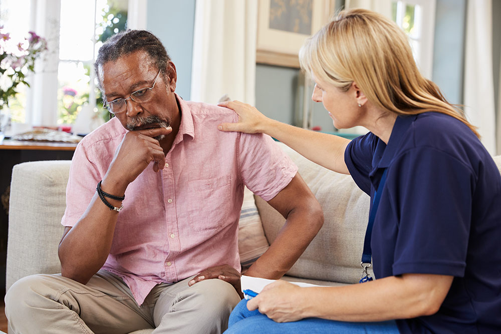 Senior man sitting on couch with caregiver, upset, worried, struggling with mental health issues in senior housing