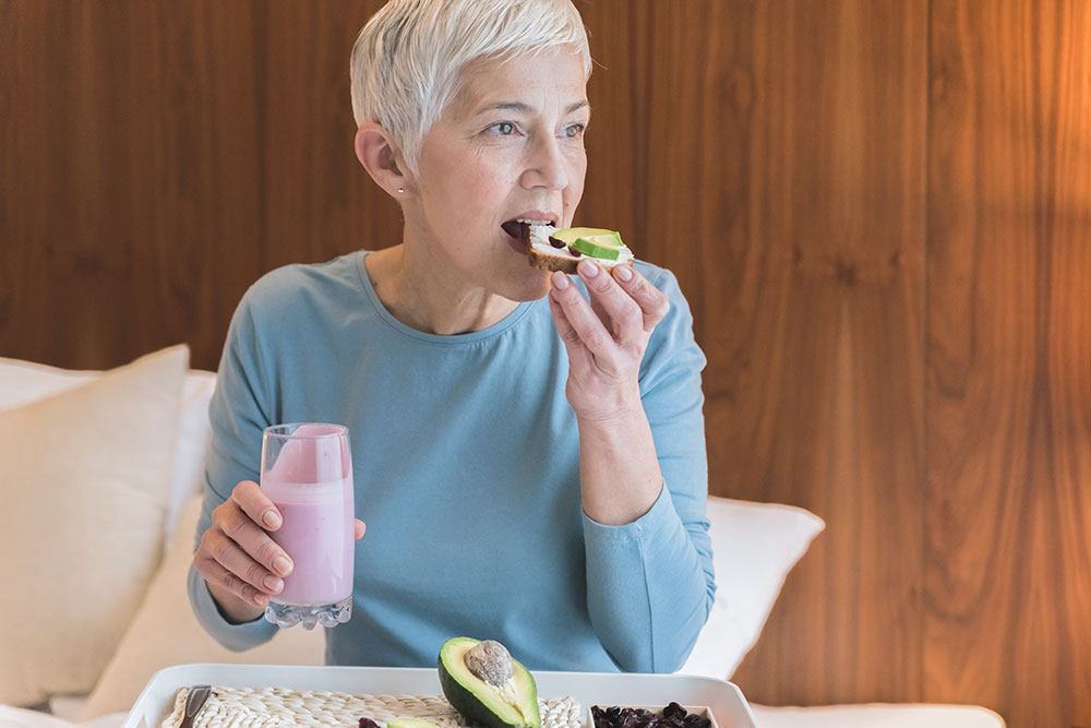 Woman sitting on couch eating avocado toast with a smoothie in the other hand