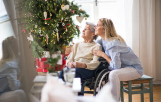 A senior woman and her adult daughter sit together in front of a holiday tree
