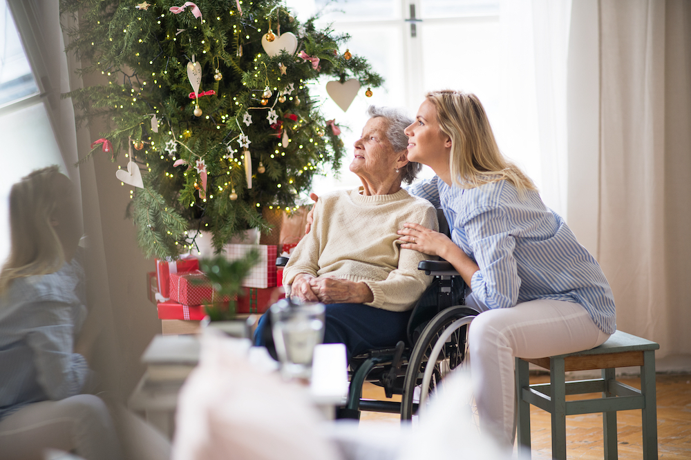 A senior woman and her adult daughter sit together in front of a holiday tree
