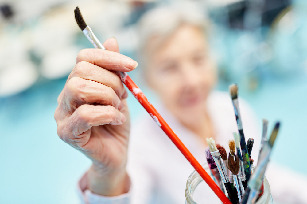 A senior woman reaching for a paintbrush while taking a painting lesson at the best memory care in Macomb, MI